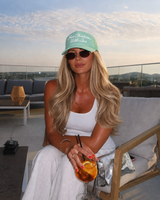 A woman sits in a lounge chair on a rooftop terrace at dusk,sunglasses, a white tank top, and white pants while holding an orange cocktail. The background shows a city skyline and hills under a cloudy sky.