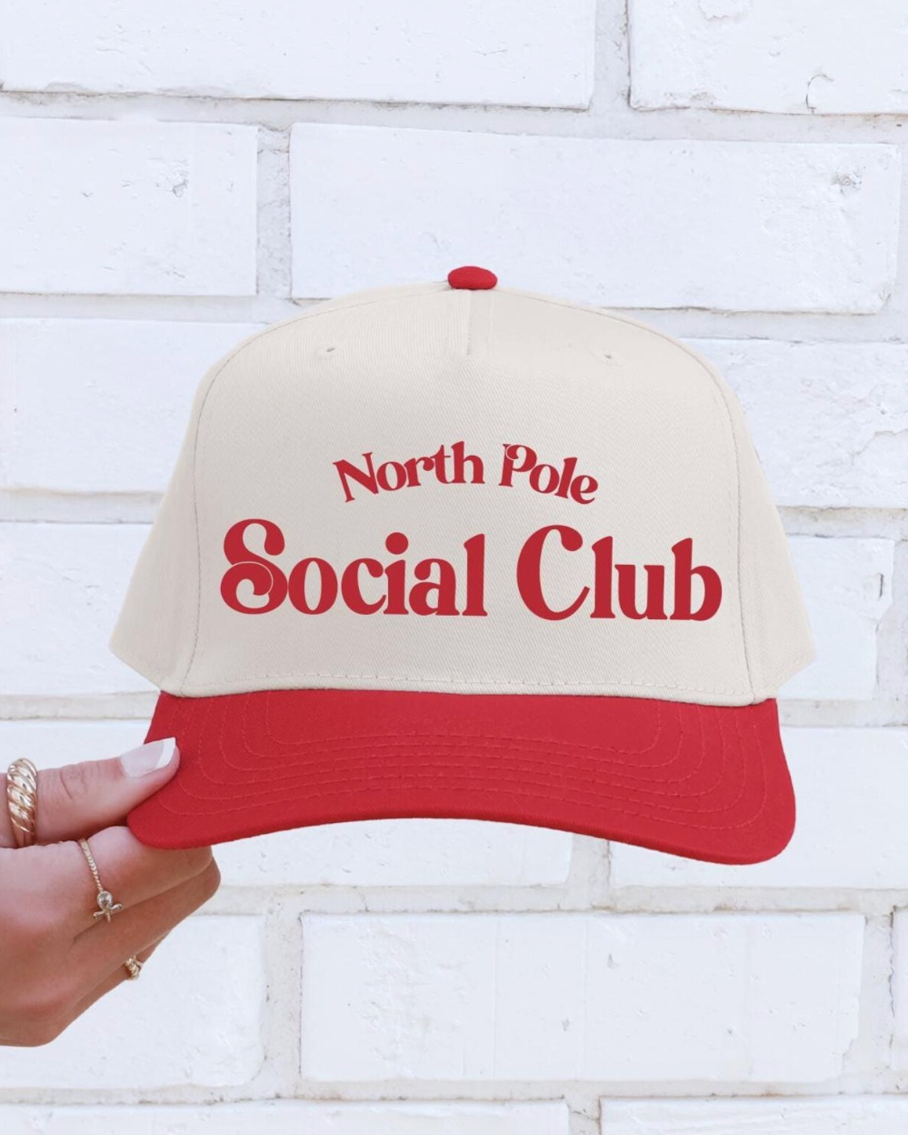 Beige and red cap with 'North Pole Social Club' text held against a white brick wall.