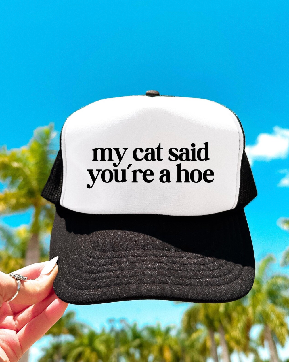 Trucker hat with text 'my cat said you're a hoe' held against a blue sky with palm trees.