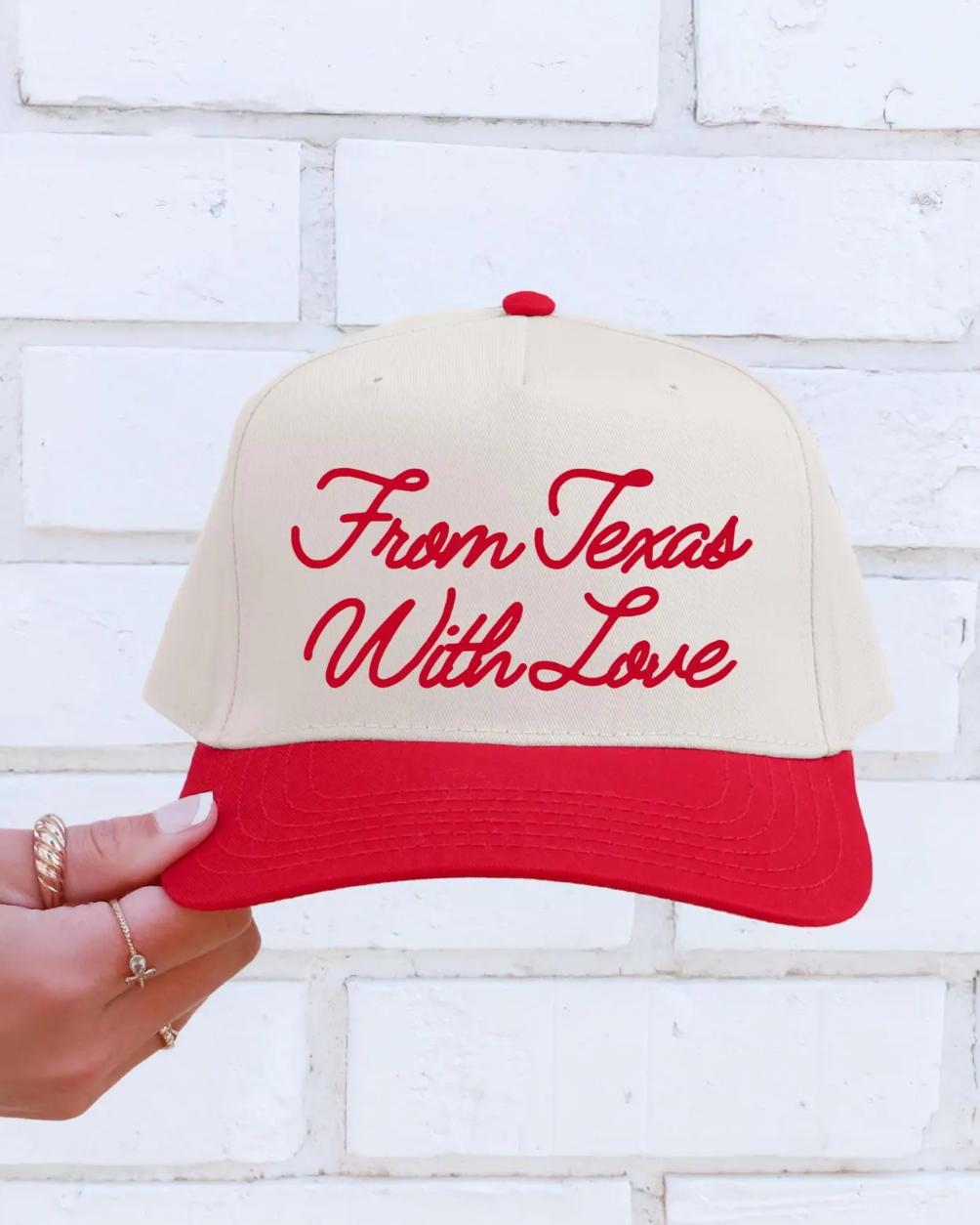 Red and beige cap with 'From Texas With Love' text held against a white brick wall.