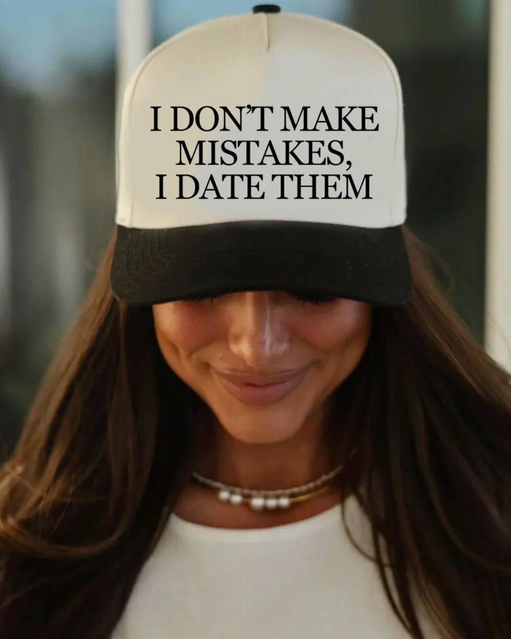 Person wearing a cap with text 'I don't make mistakes, I date them' outdoors.
