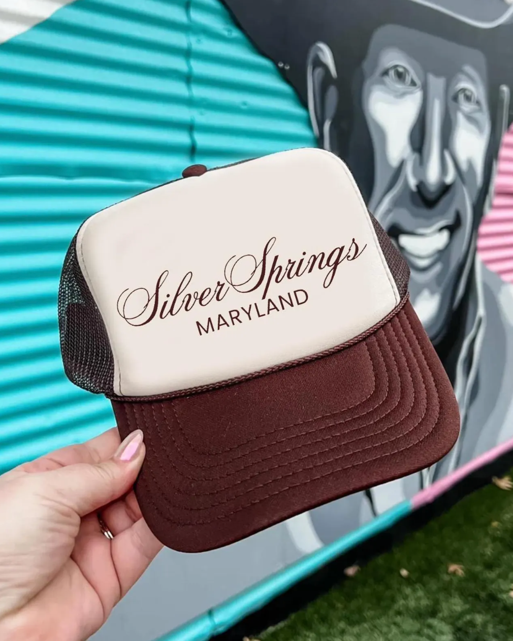 Trucker hat with 'Silver Springs Maryland' text held by a hand, with a mural of a smiling man in a cowboy hat in the background.