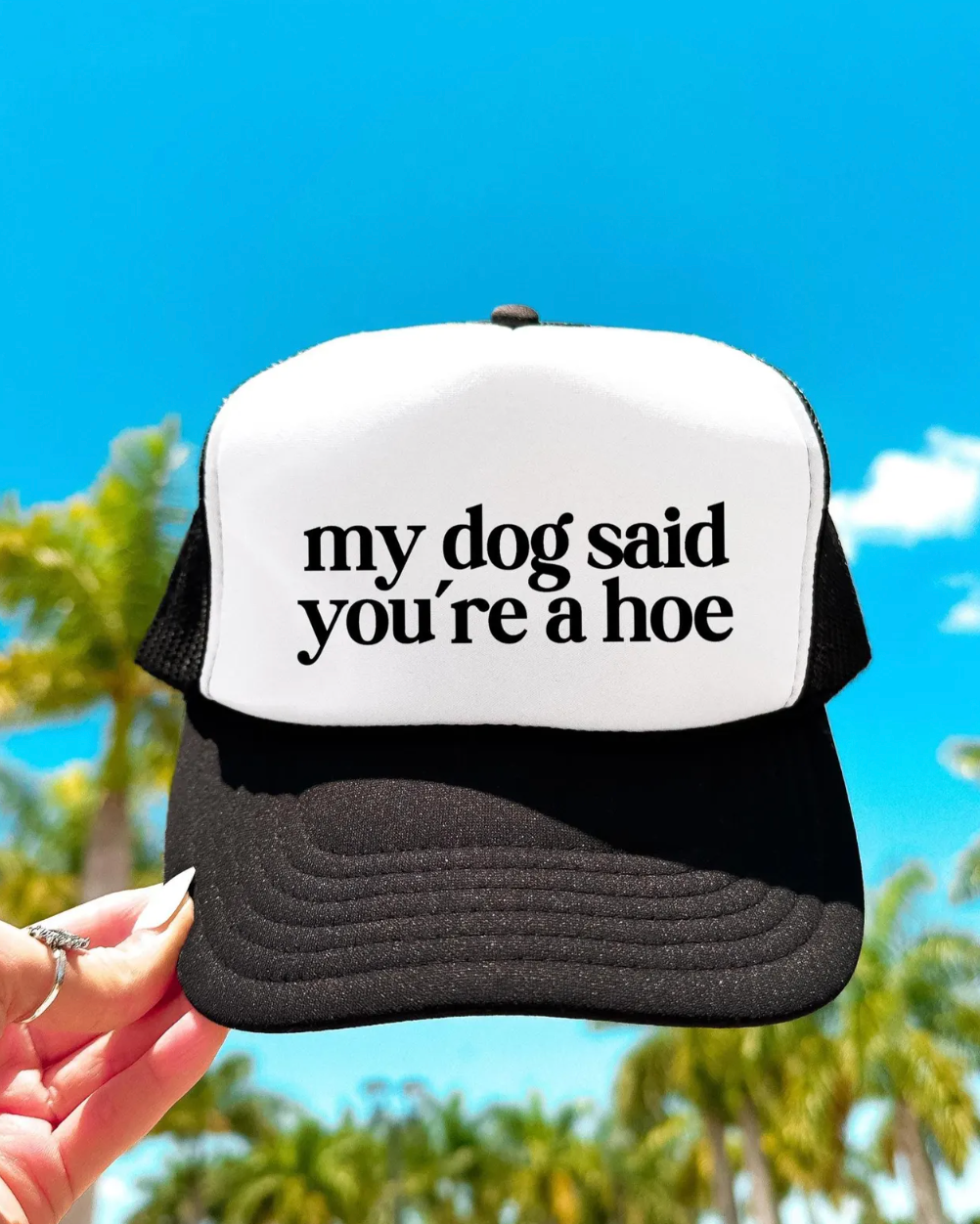 Trucker hat with text 'my dog said you're a hoe' held against a blue sky and palm trees background.