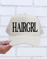Beige cap with 'HAIRGRL' text held by a hand against a white brick wall.