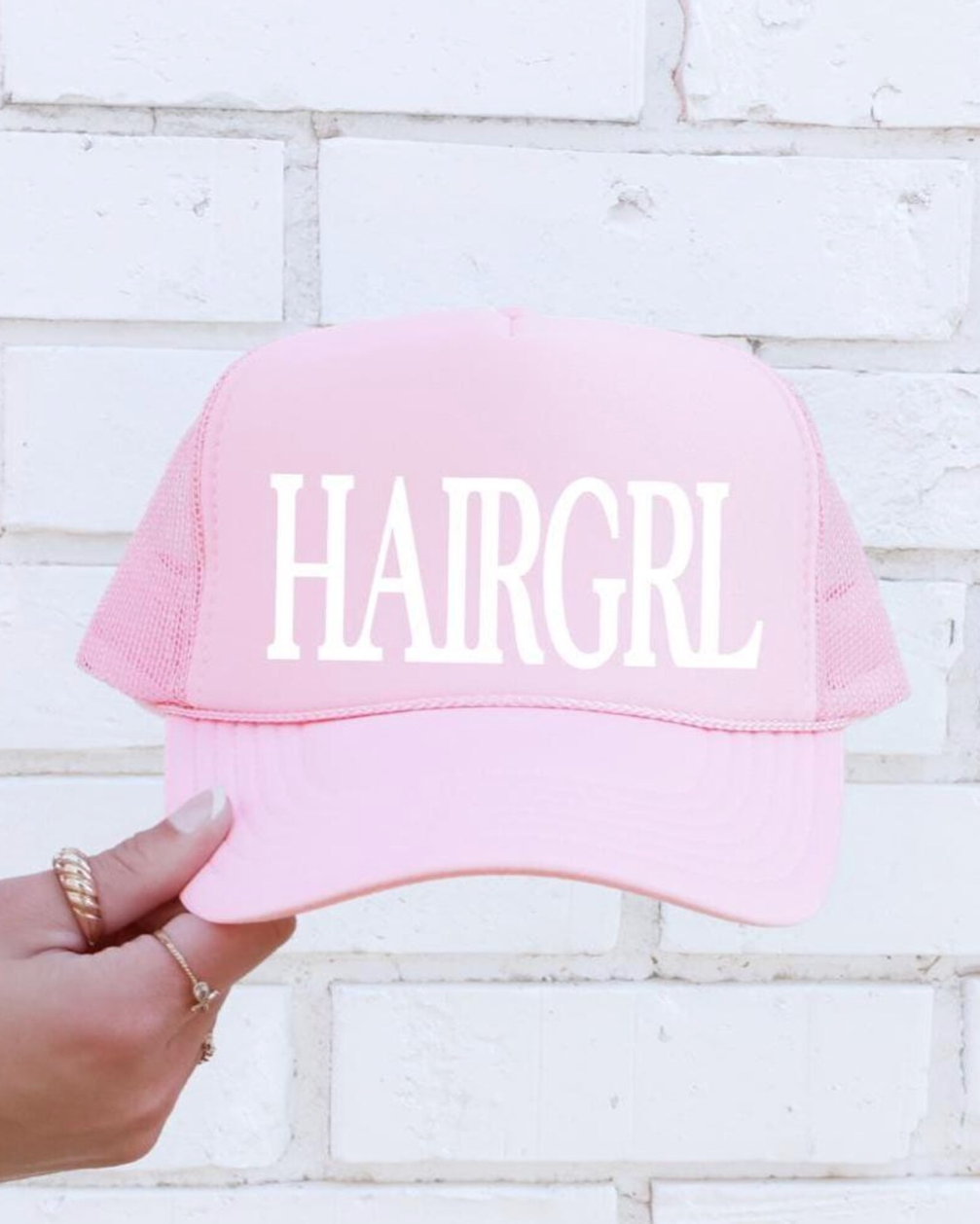 Pink cap with 'HAIRGRL' text held by a hand against a light brick wall.