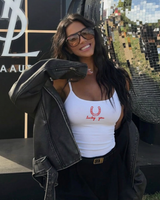 A woman wearing a white ribbed tank top with a red embroidered horseshoe and 'Lucky You' graphic, styled with a black leather jacket and black pants, posing outdoors in front of a mirrored wall.