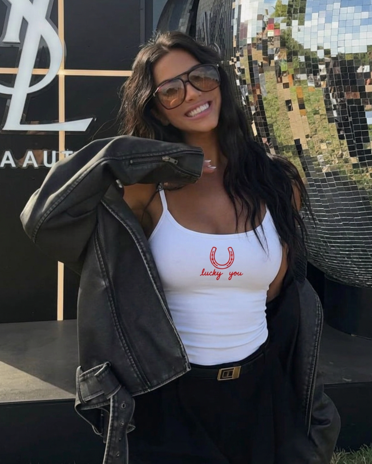 A woman wearing a white ribbed tank top with a red embroidered horseshoe and 'Lucky You' graphic, styled with a black leather jacket and black pants, posing outdoors in front of a mirrored wall.