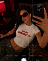 A woman wearing a white cropped t-shirt with 'MAIN CHARACTER' printed in red text, posing for a mirror selfie in a dimly lit room with a leopard-print background.