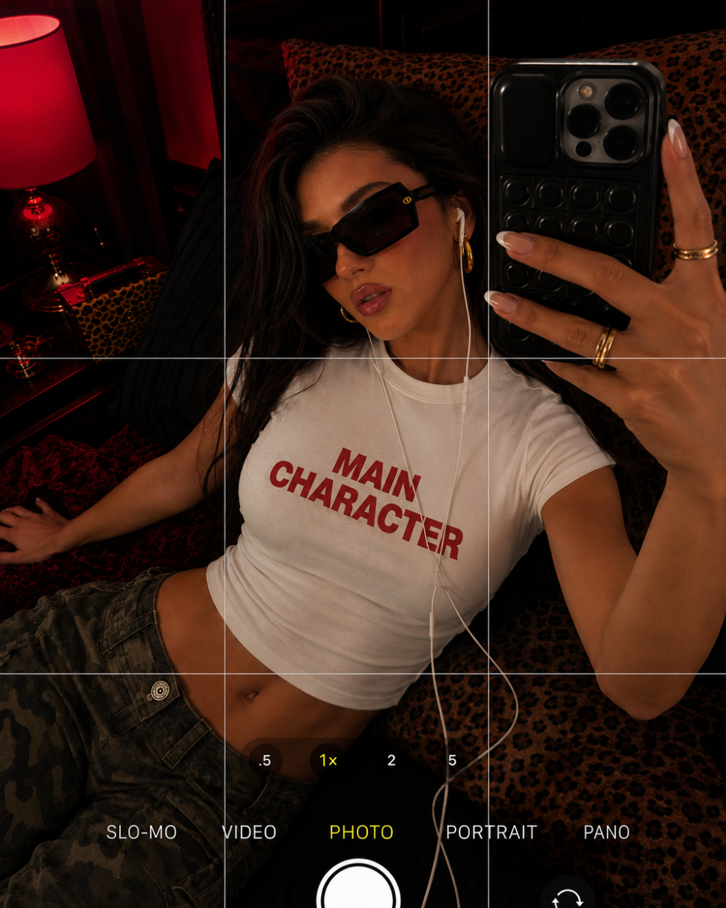 A woman wearing a white cropped t-shirt with 'MAIN CHARACTER' printed in red text, posing for a mirror selfie in a dimly lit room with a leopard-print background.