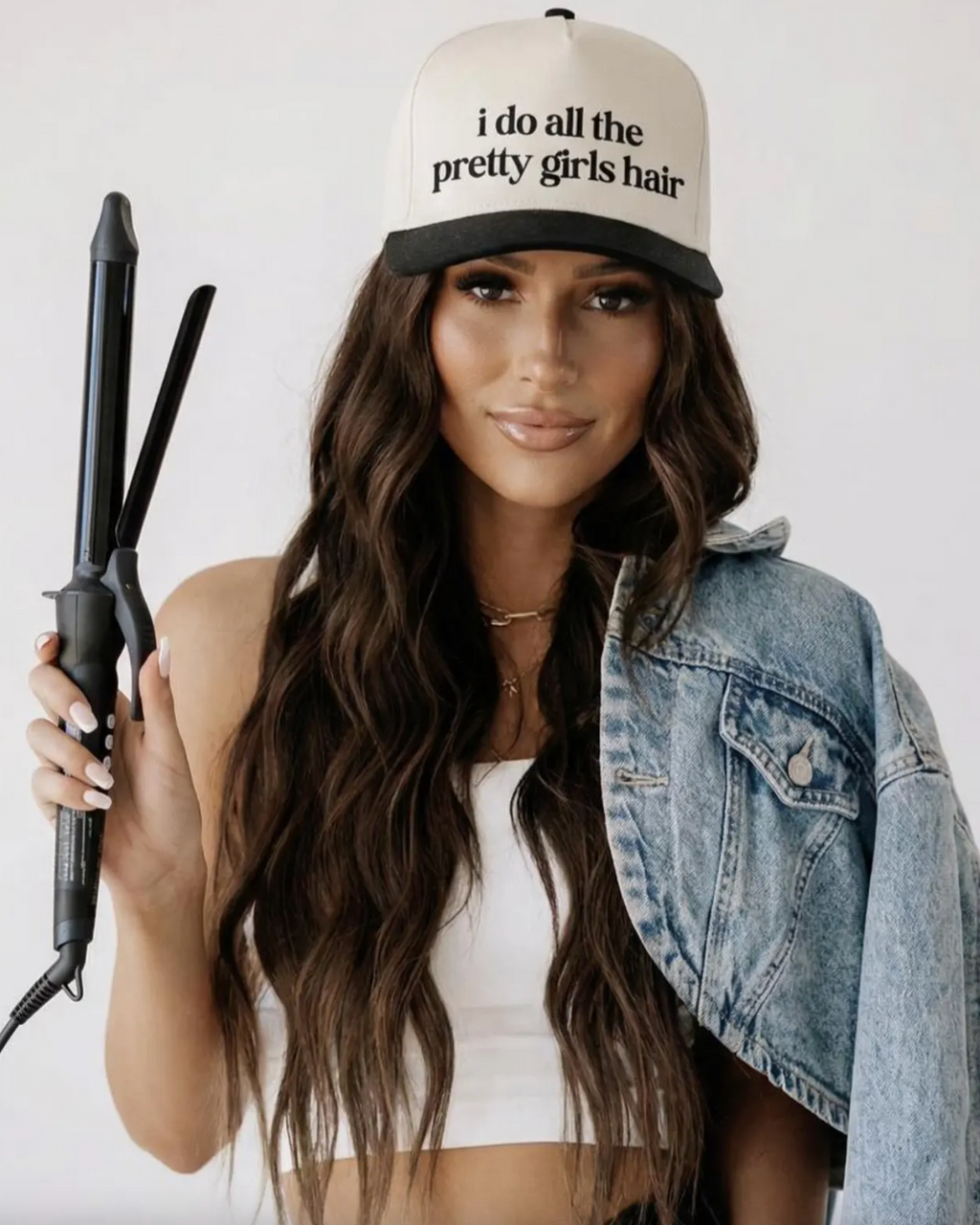 Woman holding a hair straightener wearing a cap with text, on a plain background