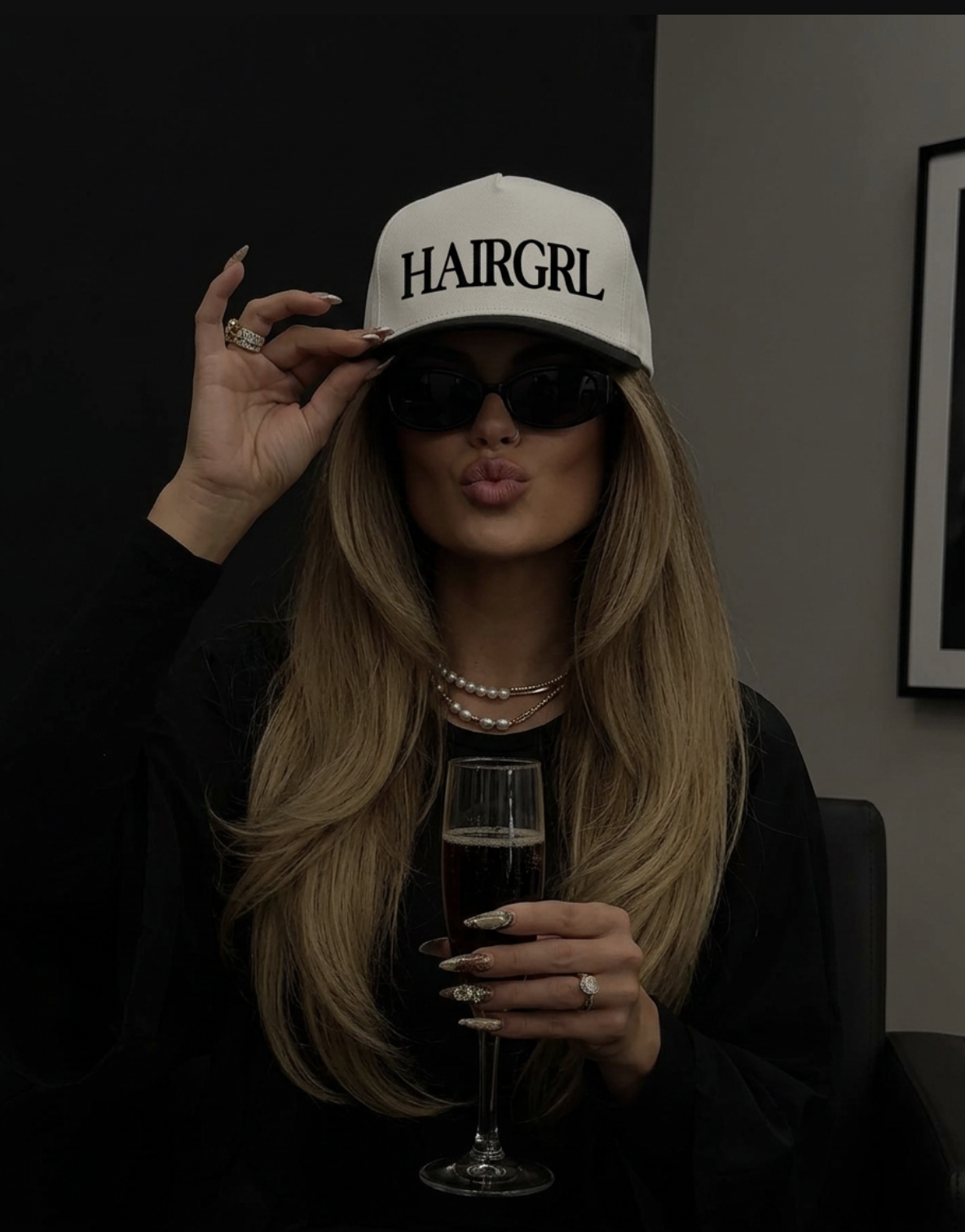 Person wearing a 'HAIRGRL' cap and holding a glass, with a dark background.
