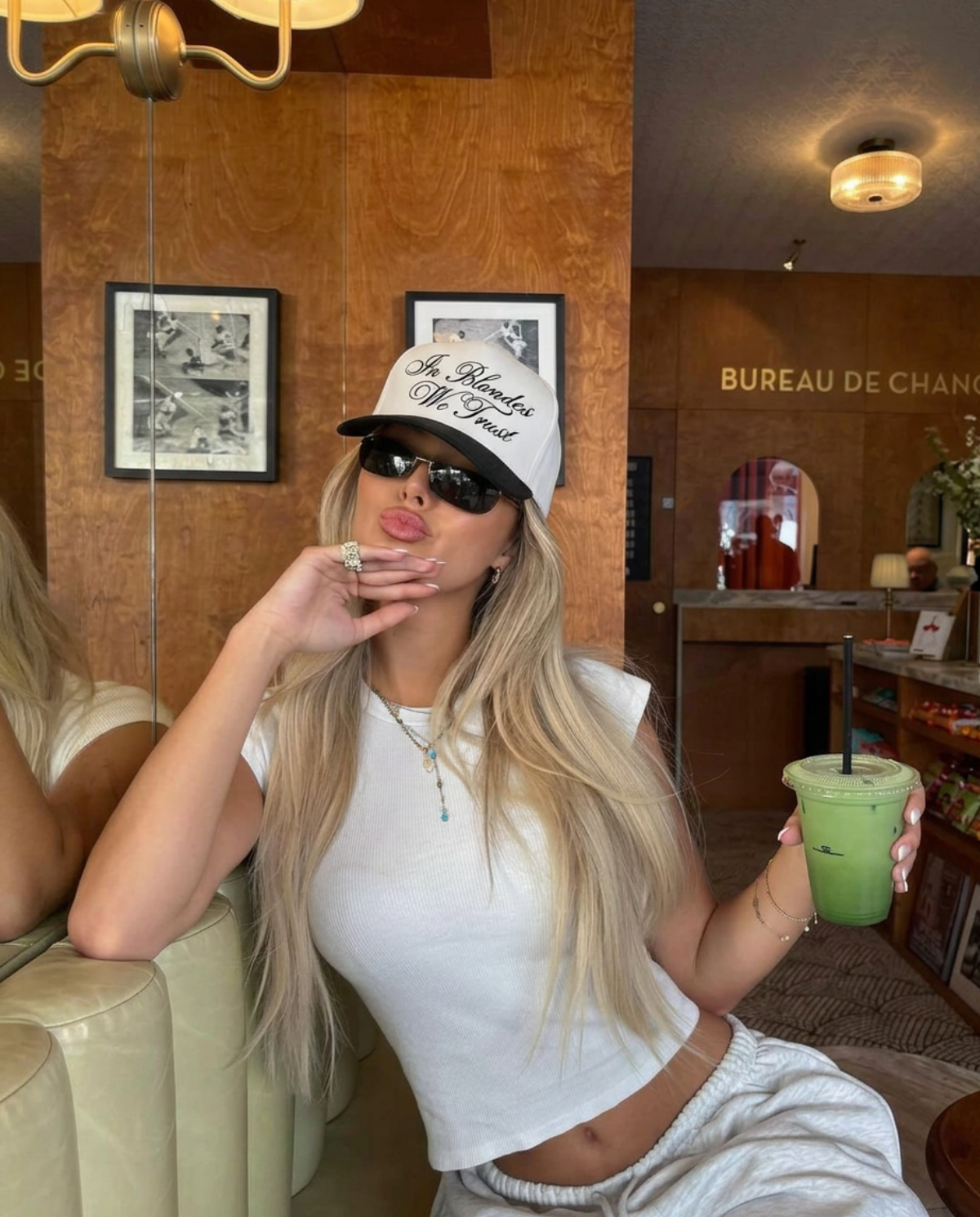 Woman in a stylish outfit with a cap and sunglasses, holding a green cup in a room with wooden walls and framed pictures.