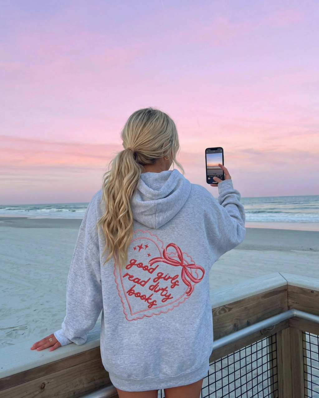 Person taking a photo of a sunset at the beach wearing a hoodie with text.