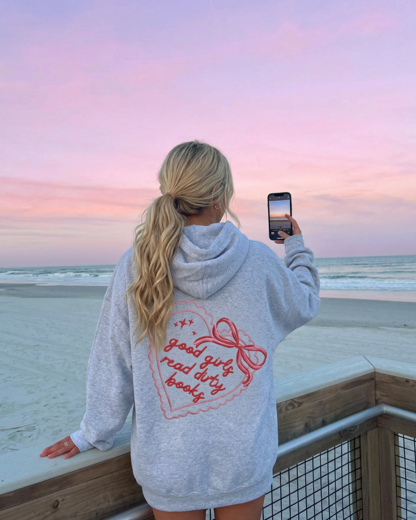 Person taking a photo of a sunset at the beach wearing a hoodie with text.