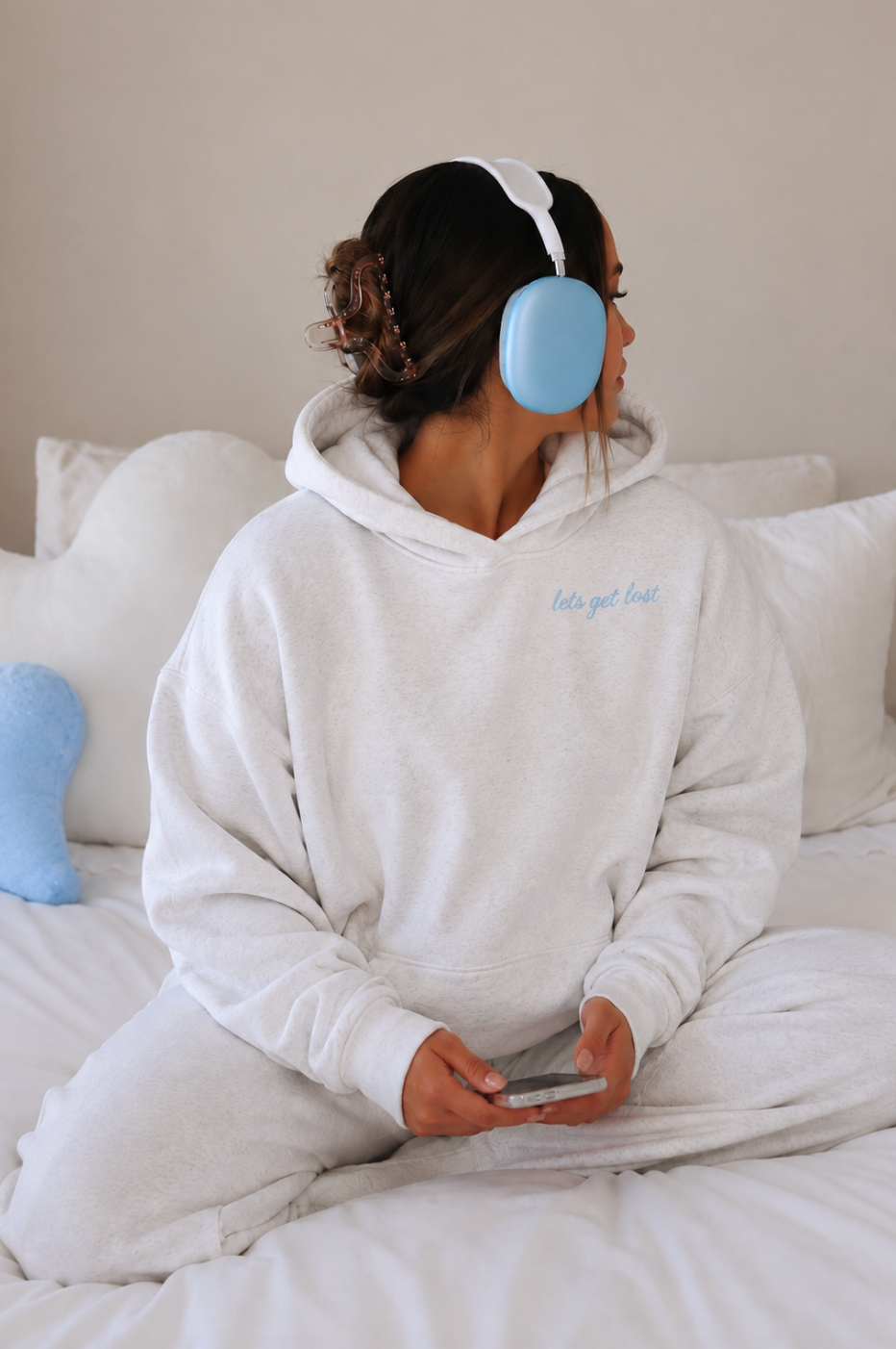 Person wearing a white hoodie with text, blue ear muffs, and holding a phone on a bed.