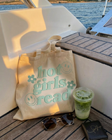 A beige canvas tote bag with the words 'hot girls read' in mint green and smiley face details, sitting on a wooden deck next to a drink, sunglasses, and a wallet on a boat.