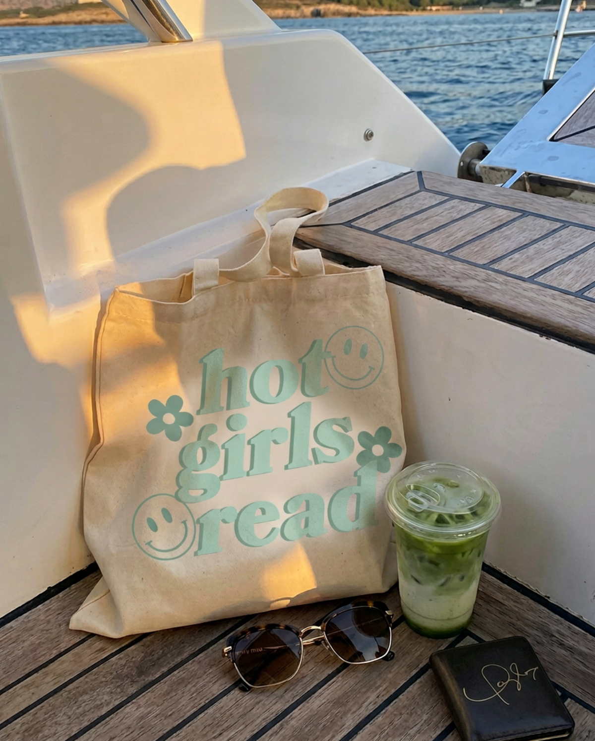 A beige canvas tote bag with the words 'hot girls read' in mint green and smiley face details, sitting on a wooden deck next to a drink, sunglasses, and a wallet on a boat.
