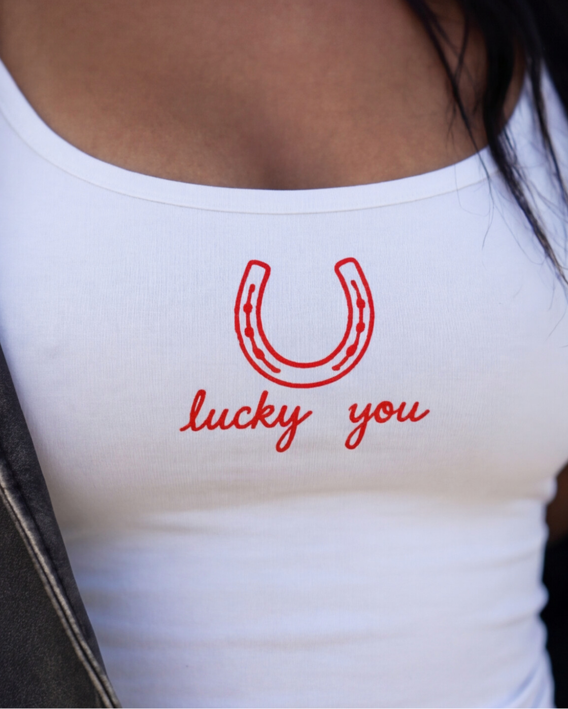 A close-up of a white ribbed tank top worn by a person, featuring a red embroidered horseshoe and the words 'lucky you' in red script on the chest.