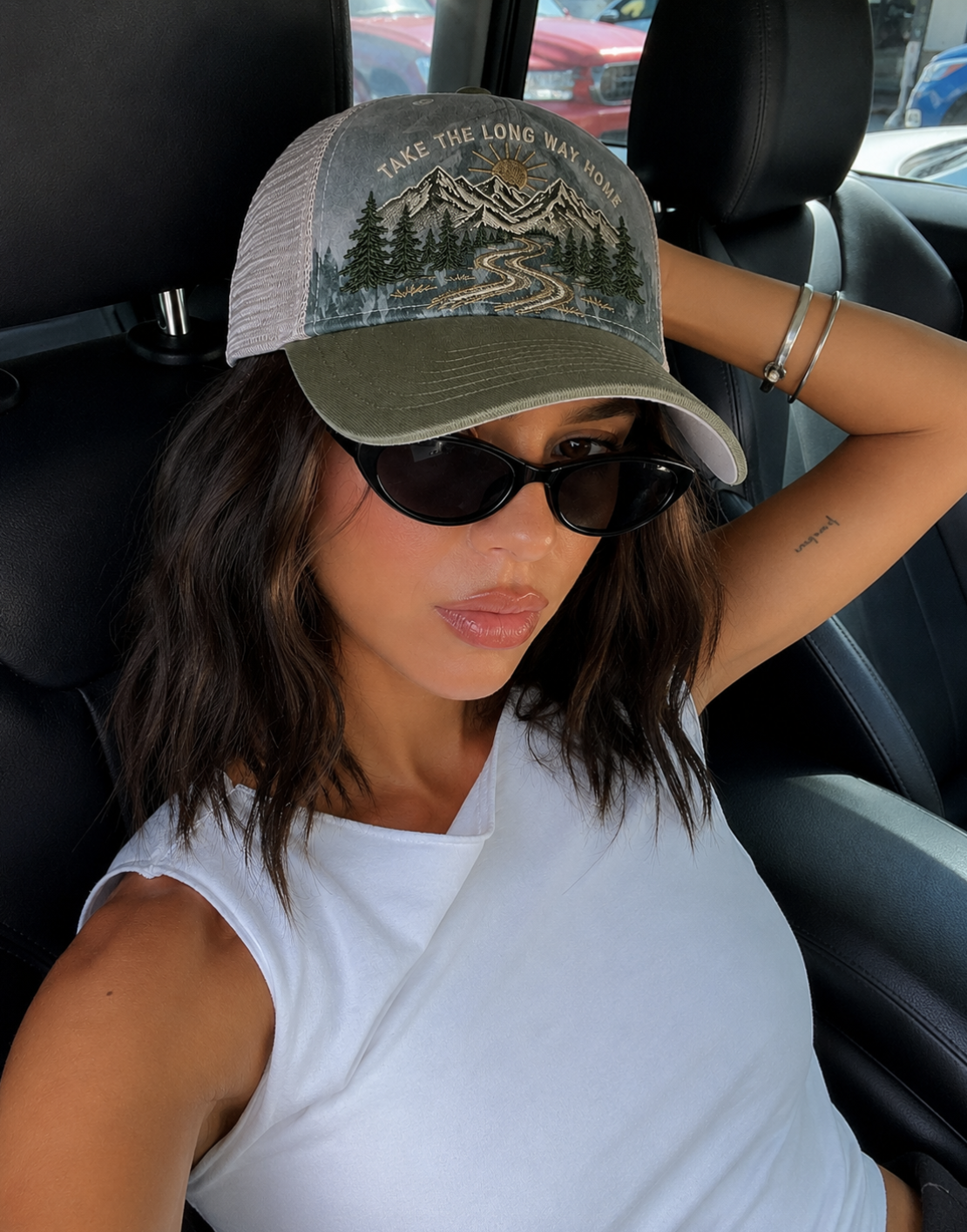 A woman sitting in a car wearing a trucker hat with a mountain and forest graphic and the text 'TAKE THE LONG WAY NORTH'.