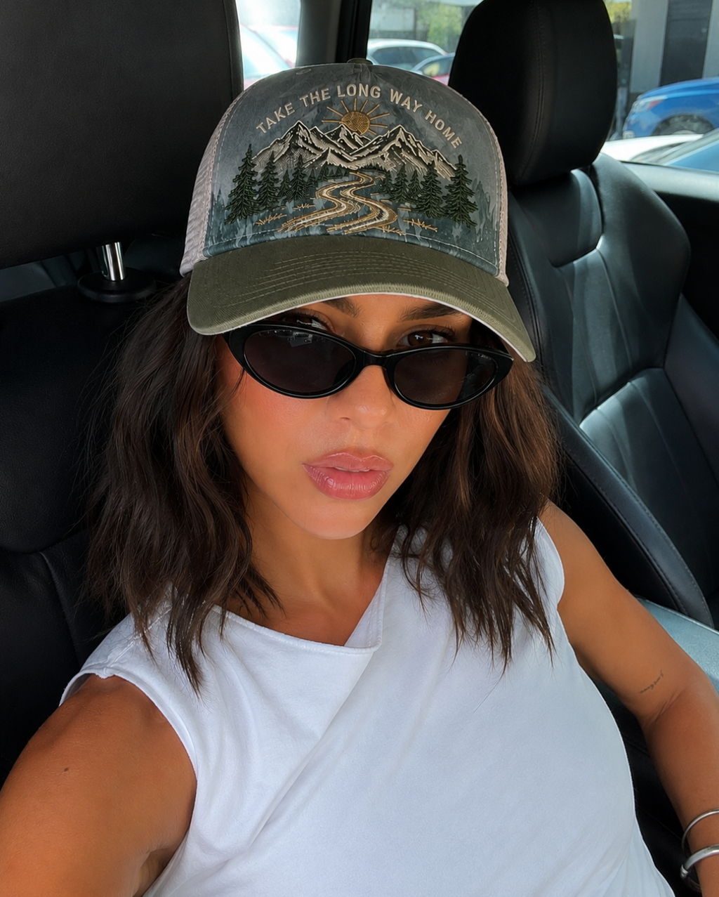 A woman wearing a trucker hat with a mountain and pine tree graphic and the text 'TAKE THE LONG WAY HOME' in a white car interior.