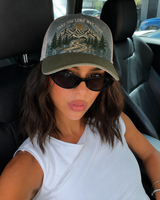 A woman wearing a trucker hat with a mountain and pine tree graphic and the text 'TAKE THE LONG WAY HOME' in a white car interior.