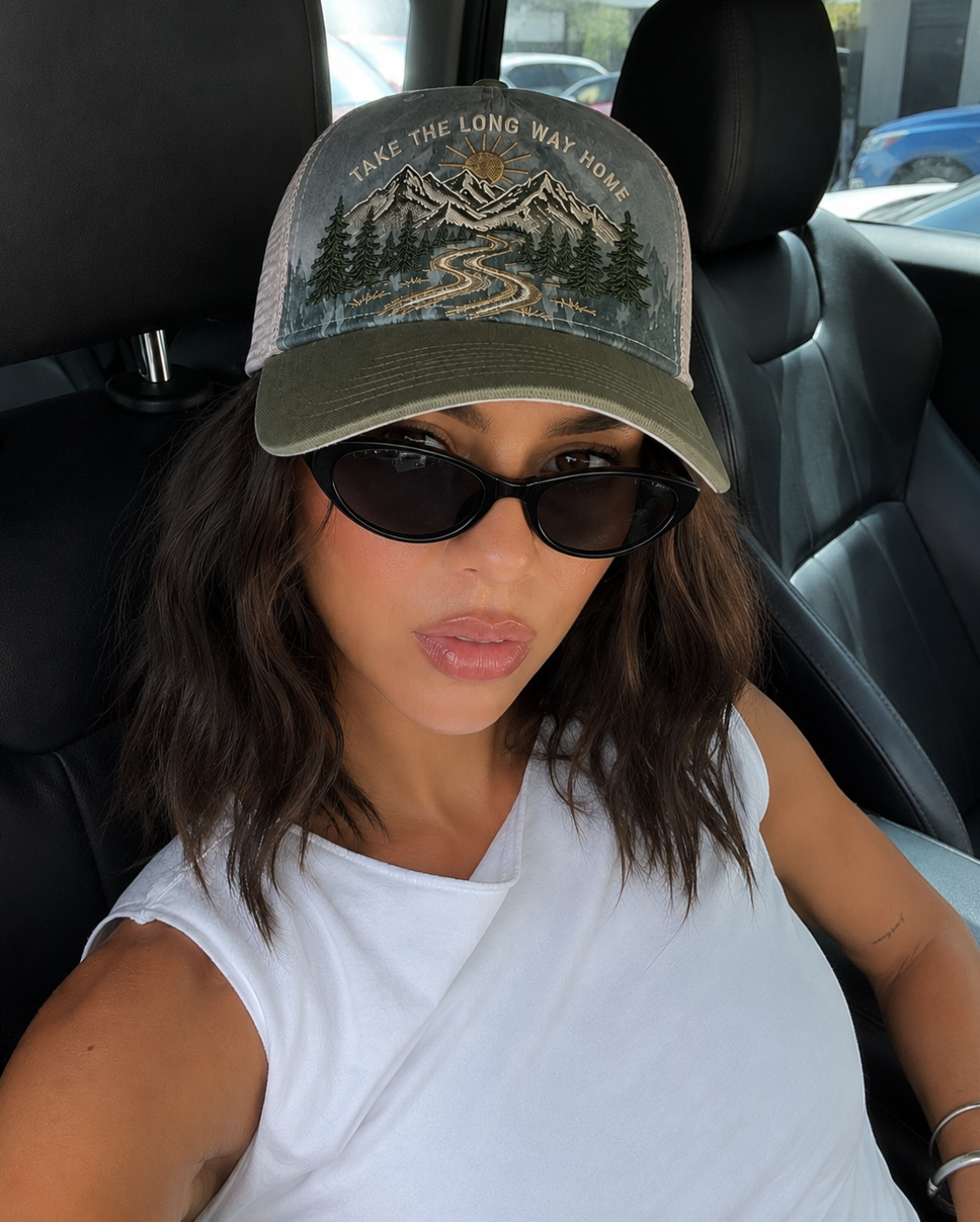 A woman wearing a trucker hat with a mountain and pine tree graphic and the text 'TAKE THE LONG WAY HOME' in a white car interior.