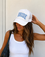 A woman wearing a white trucker-style cap with 'BRIDE' printed in blue block letters on the front, paired with a white ribbed tank top and a black shoulder bag.