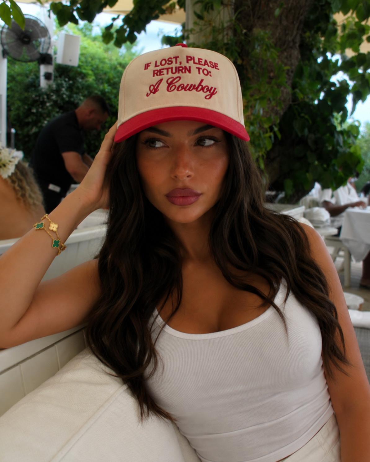 A woman wearing a cream and red trucker hat with the text 'IF LOST, PLEASE RETURN TO: A Cowboy' in red script, posing in an outdoor patio setting.