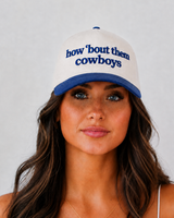 A woman wearing a white and blue baseball cap with the embroidered text 'how 'bout them cowboys' in blue on the front, photographed against a light gray background.