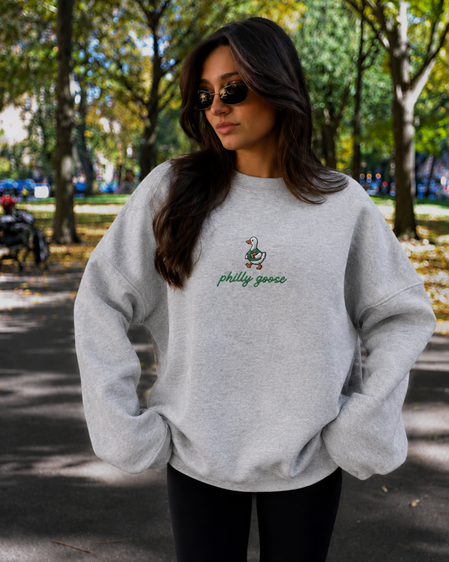 A woman wearing a light gray heather crewneck sweatshirt with a small green and white goose graphic and the text 'philly goose' on the chest, standing outdoors on a tree-lined path.
