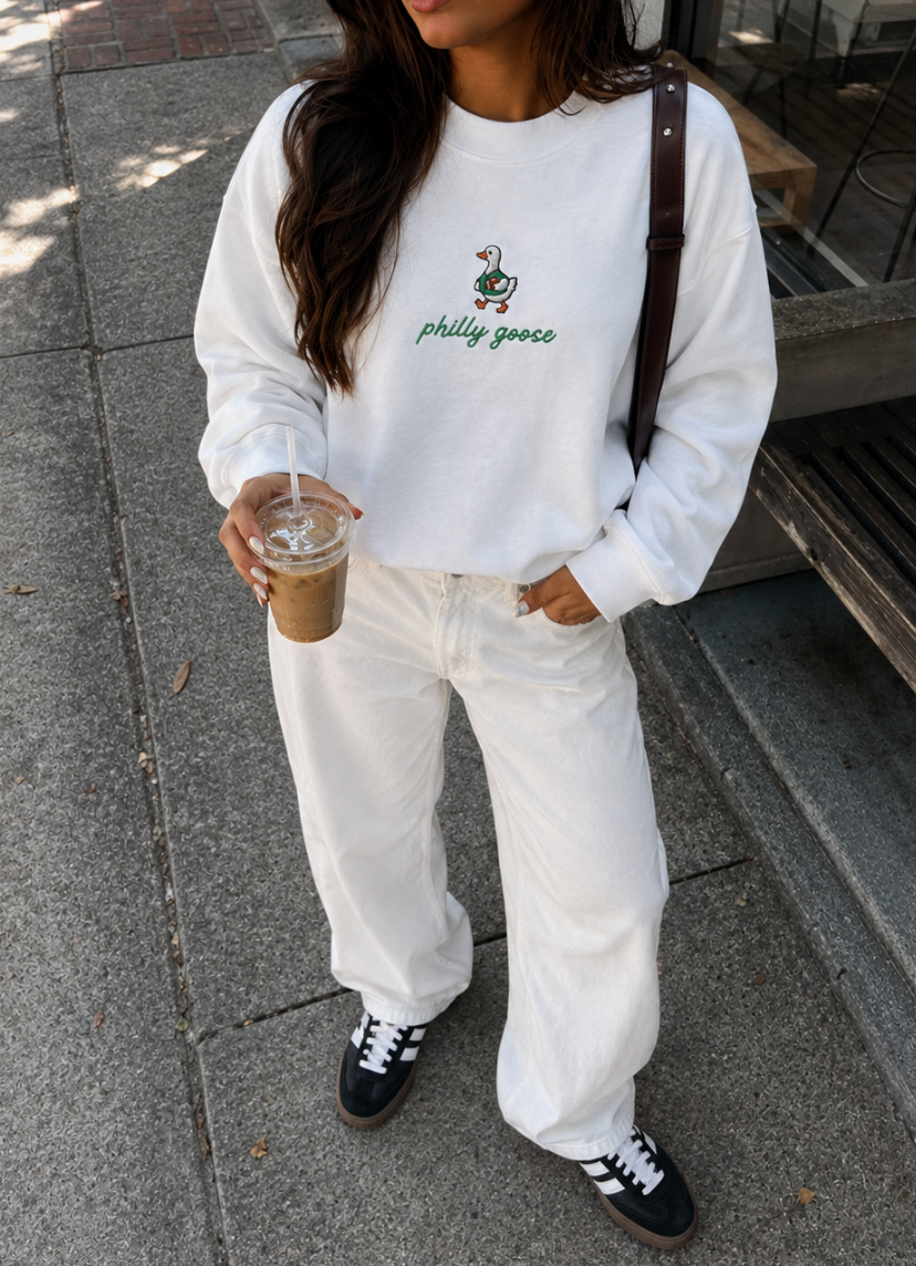 A person wearing a white crewneck sweatshirt with a green Philadelphia Phillies goose graphic and text, paired with white jeans and black sneakers, holding an iced coffee on a sidewalk.