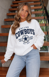 A woman wearing a white Dallas Cowboys sweatshirt and light blue jeans, posing on a wooden staircase.
