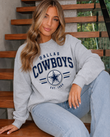 A woman sitting on wooden stairs wearing a light gray crewneck sweatshirt with 'DALLAS COWBOYS EST. 1969' printed in navy blue on the front, paired with light blue jeans.