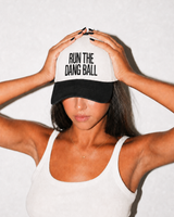 A woman wearing a white and black trucker hat with the text 'RUN THE DANG BALL' in black capital letters on the front panel, posing with her hands on the brim against a light textured wall.