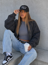 A woman sits on concrete steps wearing a black baseball cap with white text, a black bomber jacket, a gray crop top, light blue jeans, and black-and-white sneakers, while holding a white coffee cup.