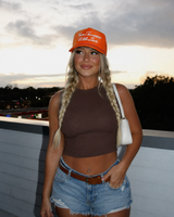 A woman posing outdoors at sunset wearing an orange Tim Excepcion 'Wild Life' baseball cap, a brown ribbed crop top, denim shorts, and carrying a white shoulder bag.