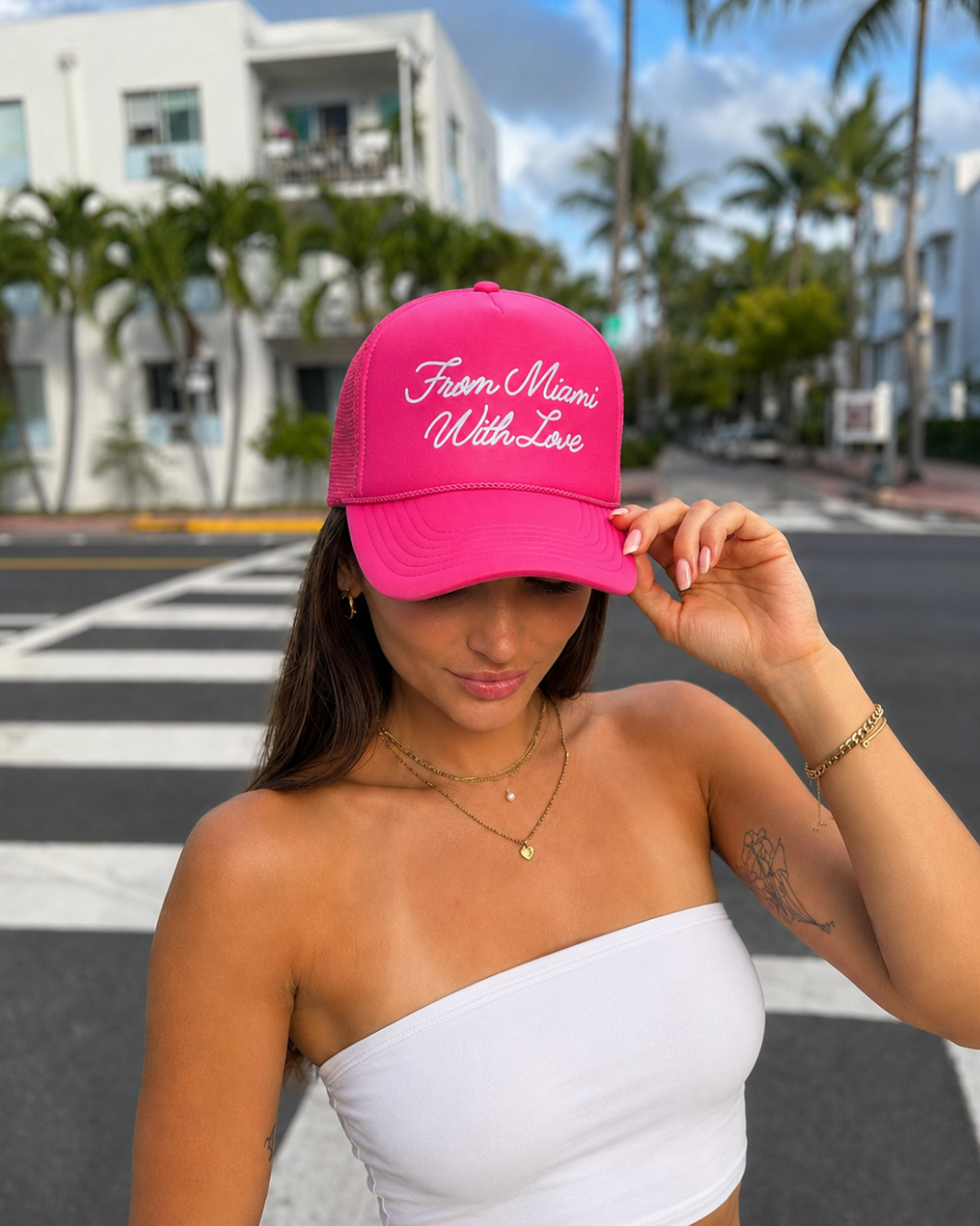 A woman wearing a bright pink trucker-style baseball cap with 'From Miami With Love' embroidered in white script on the front, photographed outdoors on a city crosswalk with palm trees and a building in the background.