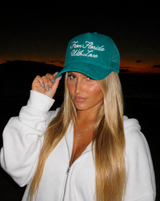 A woman wearing a teal green trucker hat with white embroidered text reading 'From Florida With Love' and a white zip-up hoodie, posing against a dark night sky with an orange horizon.