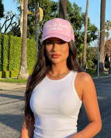 A woman wearing a light pink trucker-style baseball cap with white embroidered text reading 'Tucson, Arizona With Love' and a white ribbed tank top, standing outdoors on a sunny street lined with palm trees.