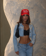 A woman posing in a light-wash denim jacket and matching jeans, wearing a red baseball cap with white text that reads 'Fan Chong United'. She is also wearing a black lace bralette and carrying a black shoulder bag, set against a textured, light-colored wall.