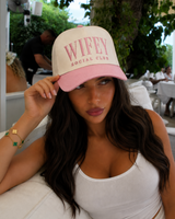 A woman wearing a beige and pink baseball cap with 'WIFETY SOCIAL CLUB' embroidered on the front, posing outdoors on a white lounge chair.