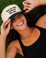 A woman lying on green grass wearing a beige trucker-style cap with black text that reads 'Hold my drink I gotta pet this dog'.