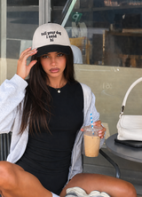 A woman sitting at a cafe wearing a black and white baseball cap with the text 'tell your dog I said hi', a black ribbed dress, a light gray zip-up hoodie, and holding an iced coffee.