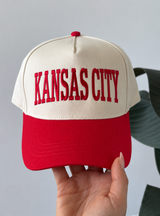 A cream and red trucker-style baseball cap with 'KANSAS CITY' embroidered in red on the front, held by a hand against a light gray background with a green leaf visible on the right.