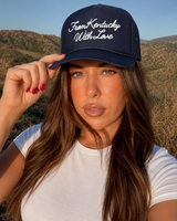 A woman wearing a navy blue trucker hat with white embroidered text reading 'From Kentucky With Love' and a white t-shirt, posing outdoors against a backdrop of hills and a blue sky.