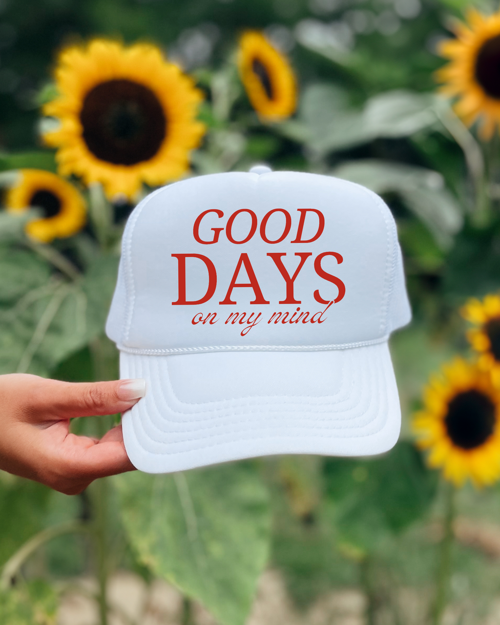 White cap with 'GOOD DAYS on my mind' text held in front of sunflowers