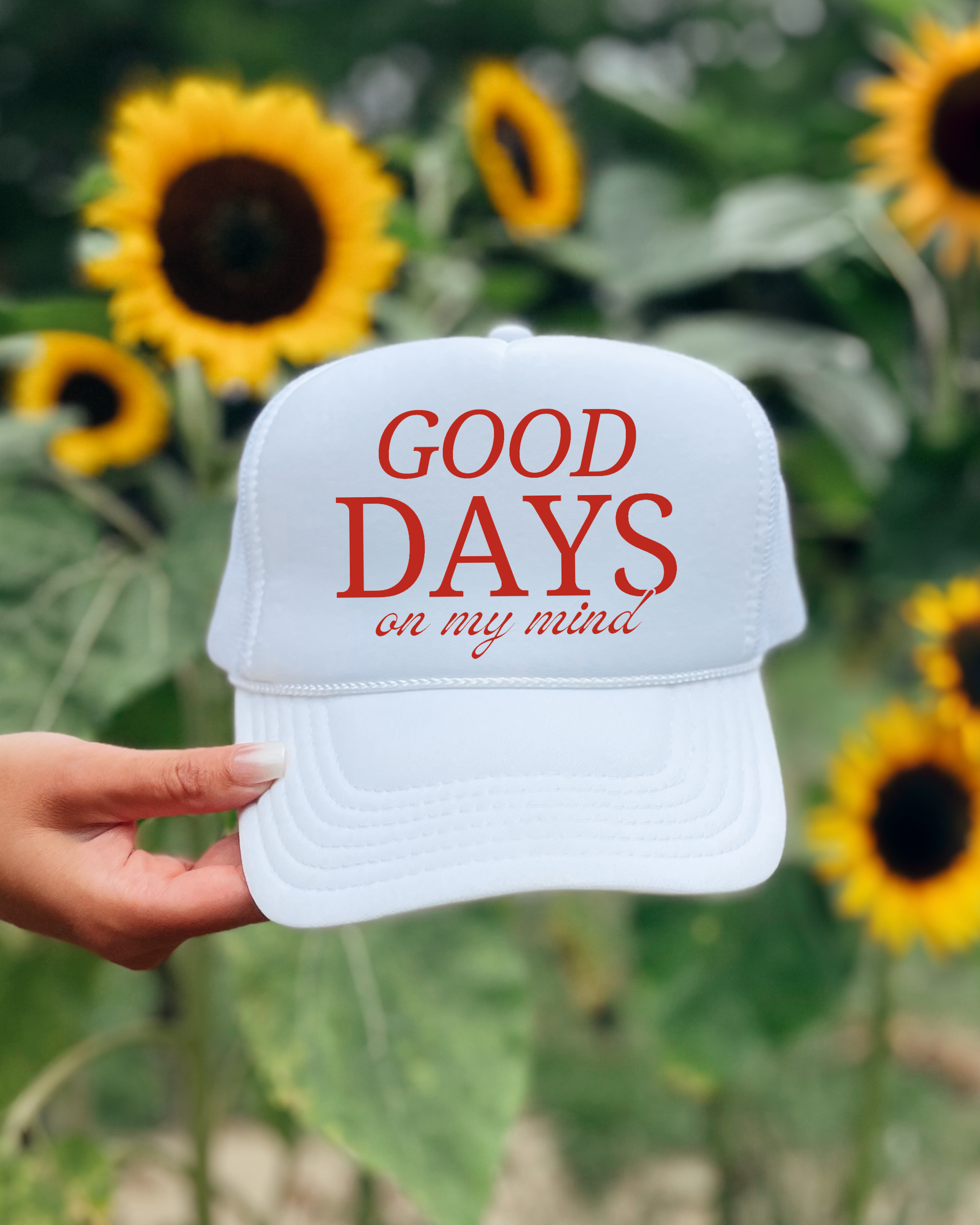 White cap with 'GOOD DAYS on my mind' text held in front of sunflowers