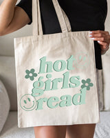 A person holding a beige canvas tote bag with the text 'hot girls read' printed in green.