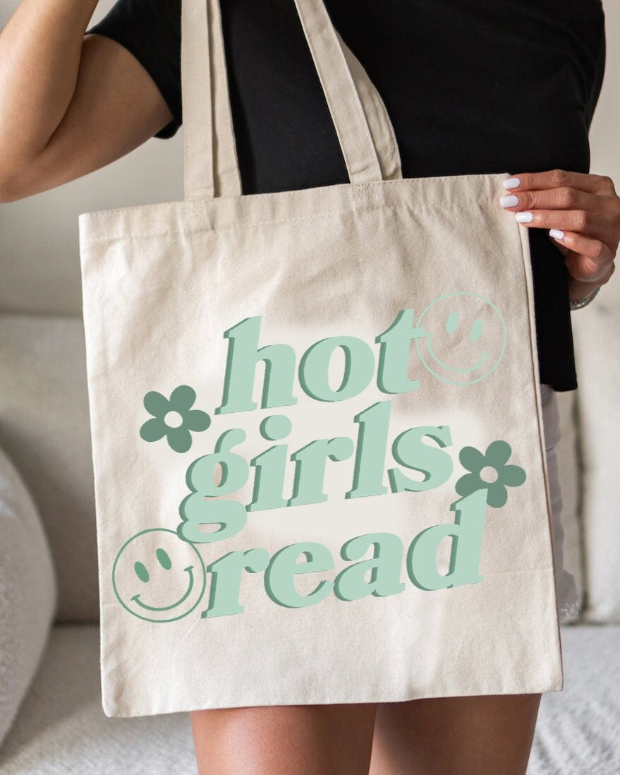 A person holding a beige canvas tote bag with the text 'hot girls read' printed in green.