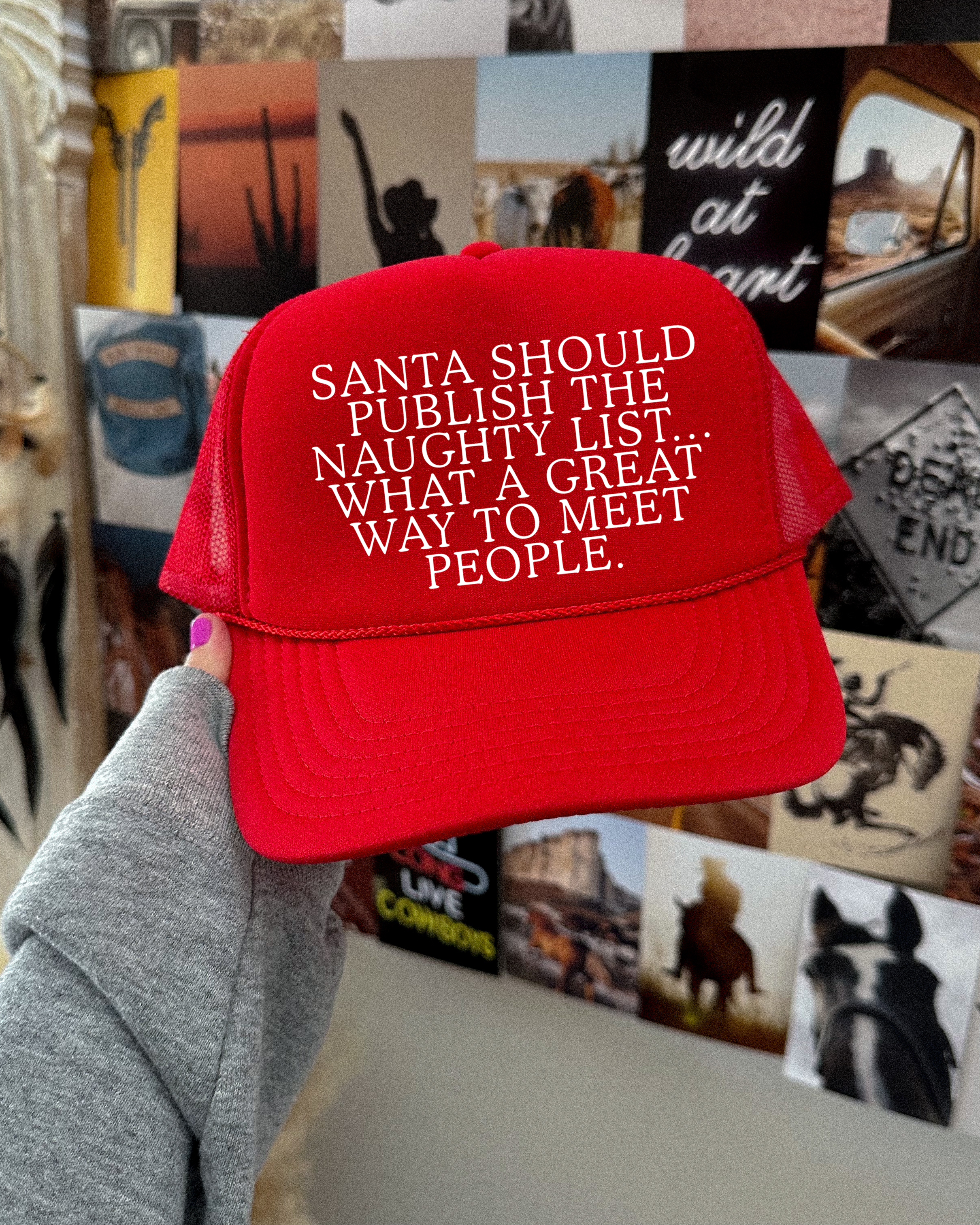 Red cap with humorous text held in front of a wall with various posters and decorations.